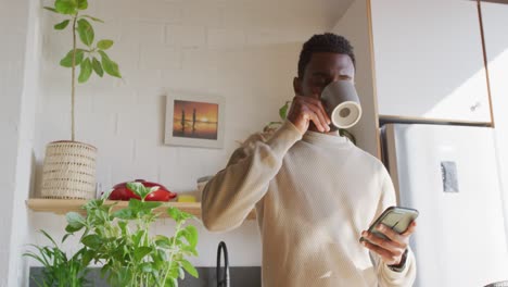 Happy-african-american-man-standing-in-kitchen,-drinking-coffee-and-using-smartphone
