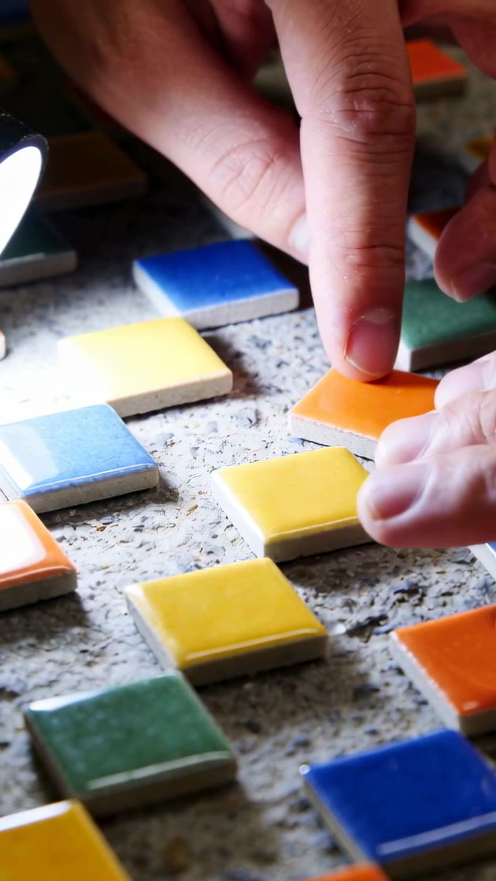 Video vertical: Mano que se extiende colocando un azulejo naranja en mortero sobre una mesa bajo una lámpara, construyendo un mosaico
