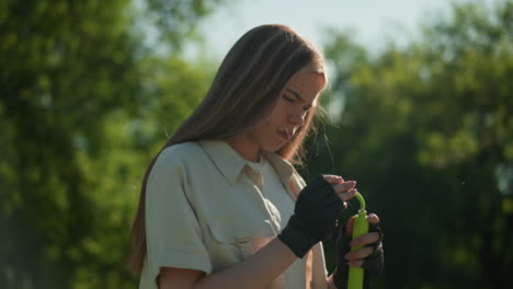 Woman with blonde hair holds green air pump in her gloved hands, inspecting it thoughtfully, against backdrop of lush green trees and natural sunlight
