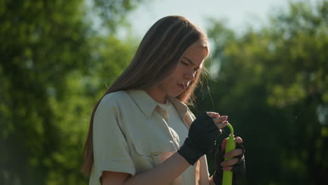 woman with blonde hair holds green air pump in her gloved hands, inspecting it thoughtfully, against backdrop of lush green trees and natural sunlight