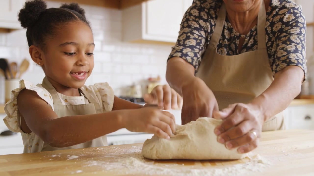Pressing adult hand, guiding child kneading dough on floured counter at home in aprons learning