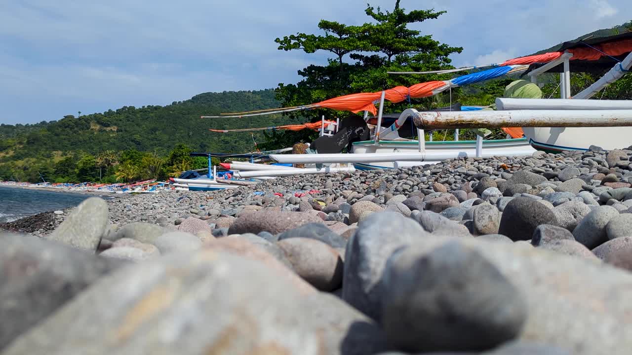 Pebble beach in Amed on tropical island of Bali Indonesia with outrigger fishing and tour boats moored along coastline