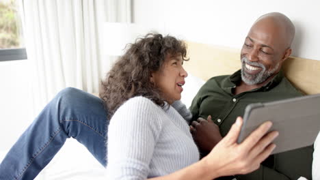 una pareja feliz, madura y diversa sentada en la cama usando una tableta y hablando en casa, en cámara lenta