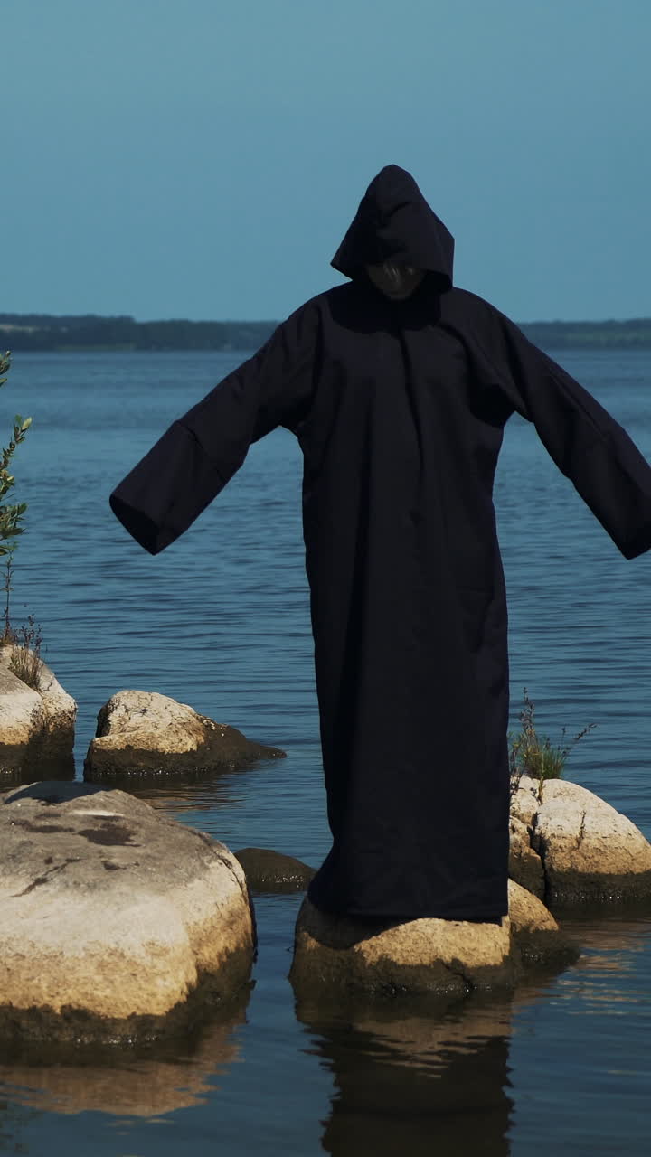 Woman devil in black cape and hood on stones near the river. Halloween costume.. Vertical video