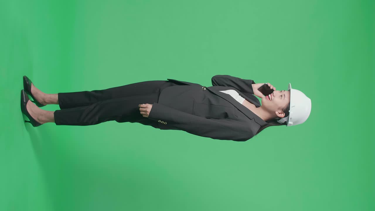 Full Body Side View Of Smiling Asian Female Engineer Celebrating While Talking On Smartphone In The Green Screen Studio