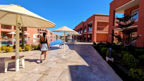 Mujer-Caminando-En-Un-Resort-De-Lujo-Con-Sombrillas-Y-Piscina,-Soleado-Telón-De-Fondo-Egipcio,-Cielo-Azul-Claro