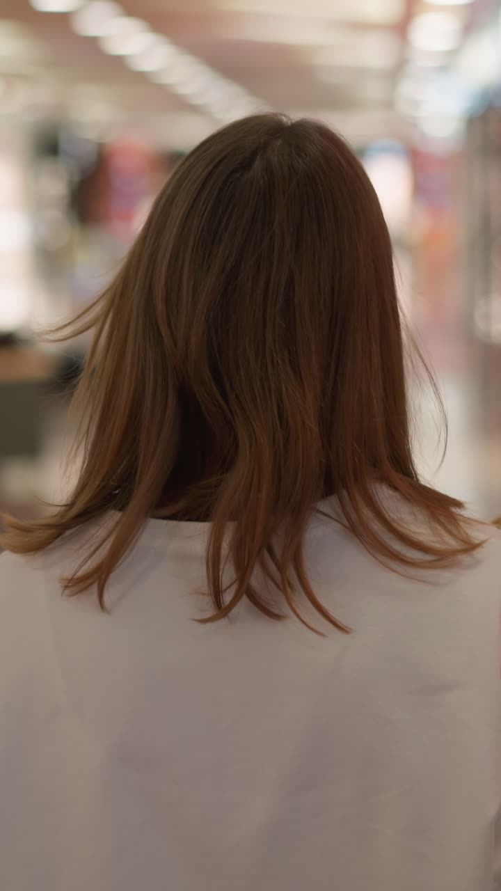 반사 유리 표면을 특징으로하는 흐릿한 배경으로 다채로운 불빛으로 조명 된 쇼핑몰을 걸어가는 색 을 입은 여성의 뒷면