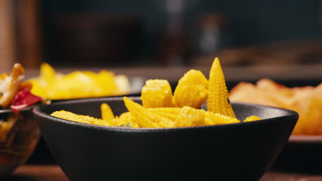 Close-up of fermented yellow mini corn with black pepper on black plate. Preservation of vegetables in glass jars. Fermentation preserved mini corns with spices.
