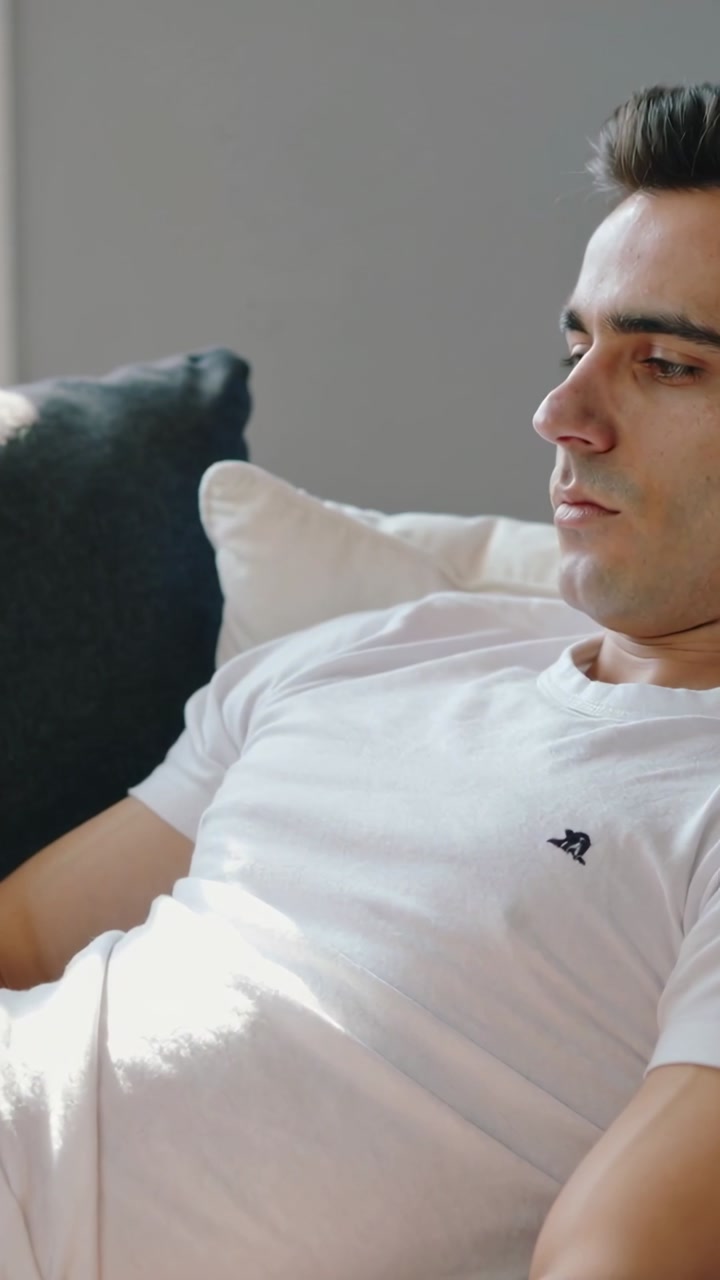 Vertical video: Showing reclining man gazing right observing offscreen on couch at home, logo shirt