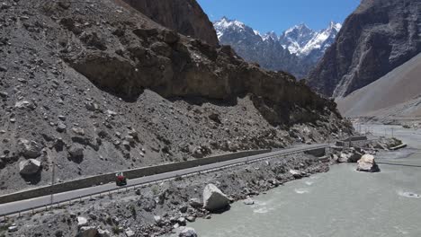 Luftaufnahme-Einer-Rikscha-Auf-Einer-Landstraße-Im-Pakistanischen-Hochland,-Die-Sich-Am-Fluss-Unter-Schneebedeckten-Berggipfeln-Entlang-Bewegt