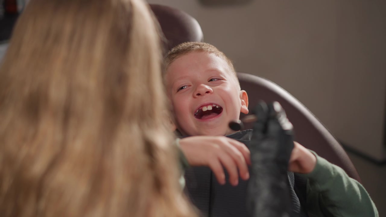 Primo piano di un bambino seduto sulla poltrona del dentista che indossa un bavaglino protettivo mentre il dentista gli esamina i denti. Il bambino interagisce con una donna adulta mostrando espressioni e movimenti delle mani.