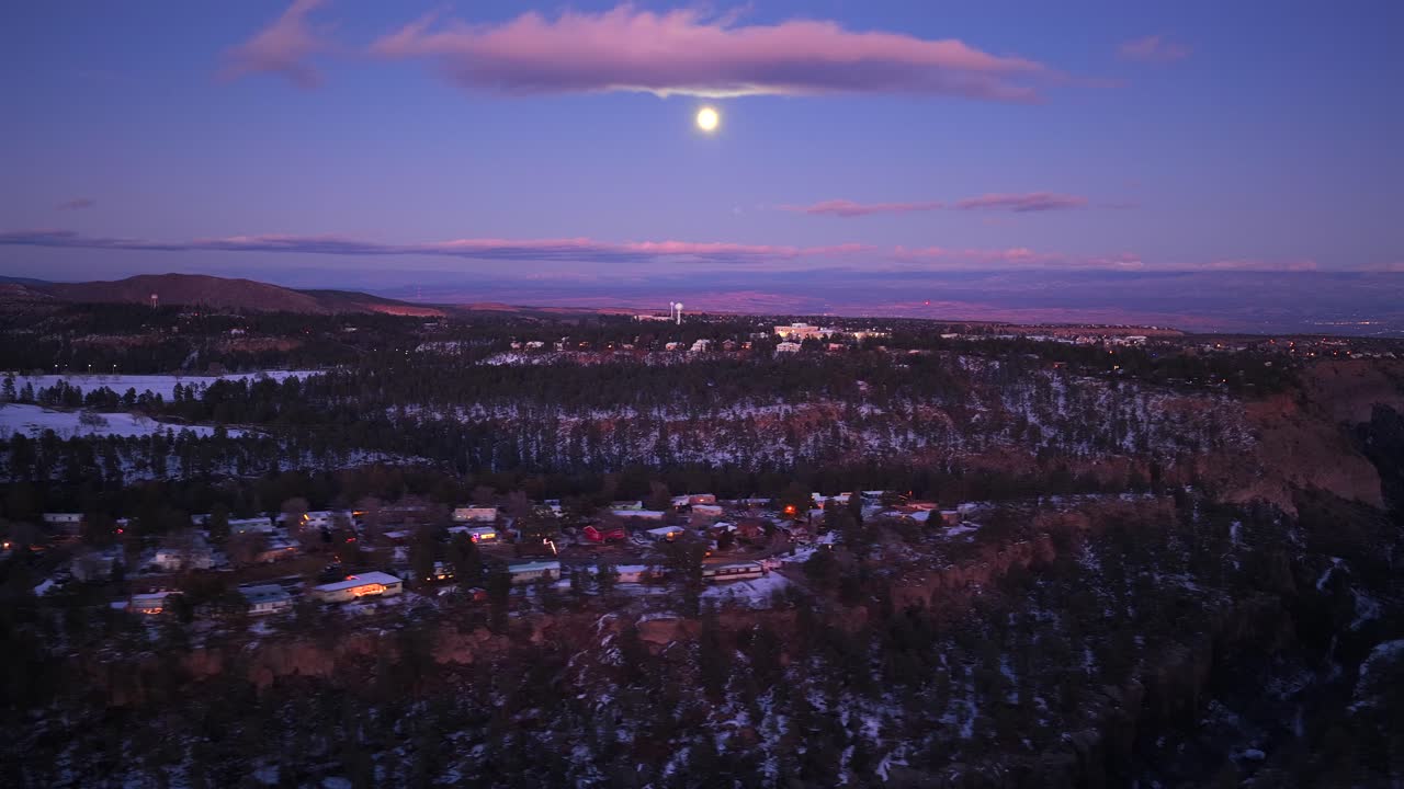 En lateral luftdias fanger en glødende fuldmåne over en alpin kløftby under den blå time. Bløde, lyserøde skyer driver over det sneklædte boliglandskab og bjergets kant i denne vinterlige tusmørke.