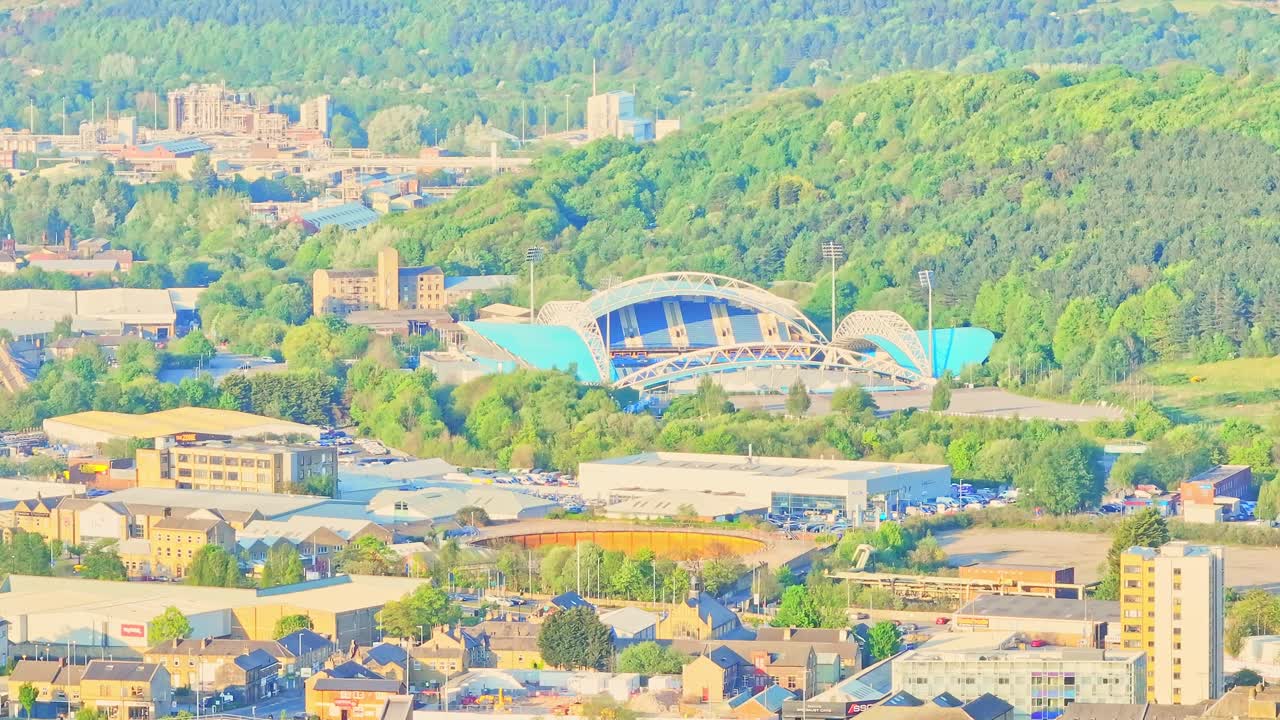 Huddersfield Town A.F.C. Stadium complex with curved roofs and training fields, surrounded by dense woodland and city structures, clear spring afternoon, aerial view, real time
