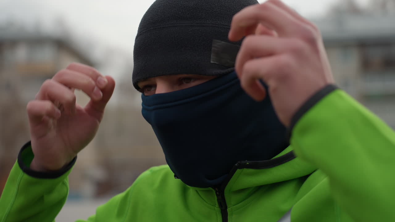 Hombre caucásico ajustándose un pasamontañas en una calle de la ciudad, con chaqueta verde y gorro, bloques de apartamentos, cielo gris, manos ajustando la tela, expresión decidida, aliento frío preparándose para correr al aire libre.