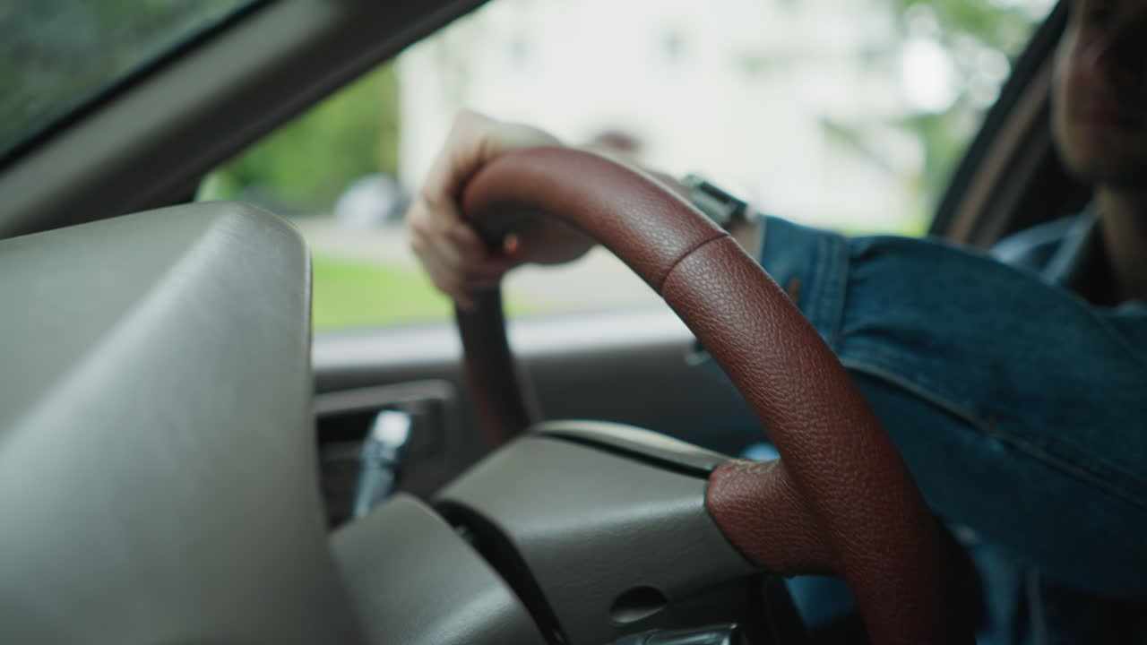 Homme tenant un volant en cuir marron à l’intérieur d’une voiture, arbres et autoroute visibles à travers la fenêtre, manche en denim et montre-bracelet dans le cadre, gros plan sur une main effectuant un virage en douceur avec une posture assurée