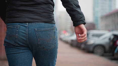 lower view of young man wearing blue jeans and black winter coat walking along urban sidewalk with parked cars and buildings in background while pedestrians stroll past on pavement during winter day