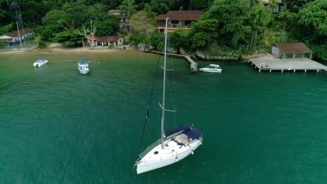 Aerial-drone-orbiting-shot-of-moored-sail-boat-near-pier-of-a-small-beach-with-one-house-and-just-a-few-boats