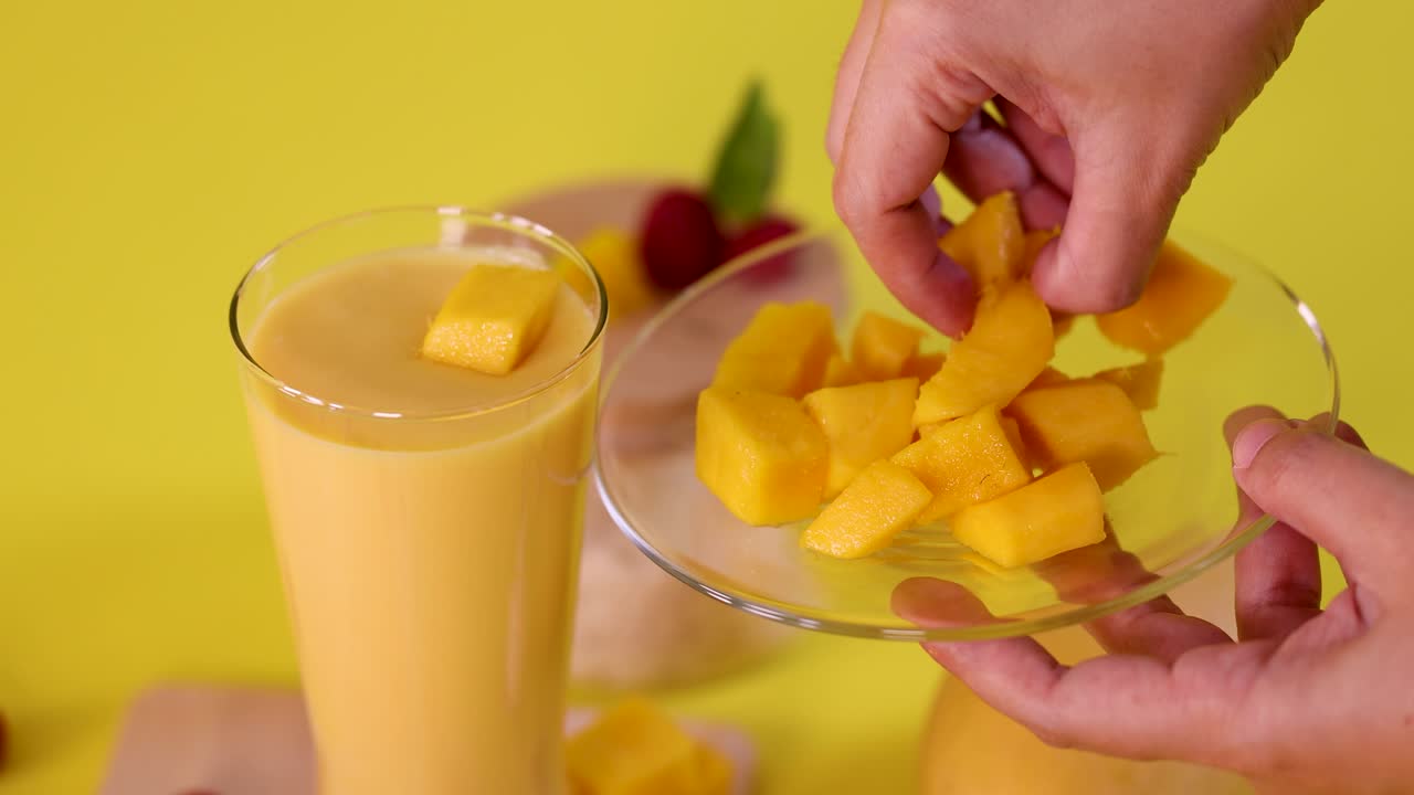 Hand places mango cubes into smoothie glass on yellow background, bright lighting, static camera