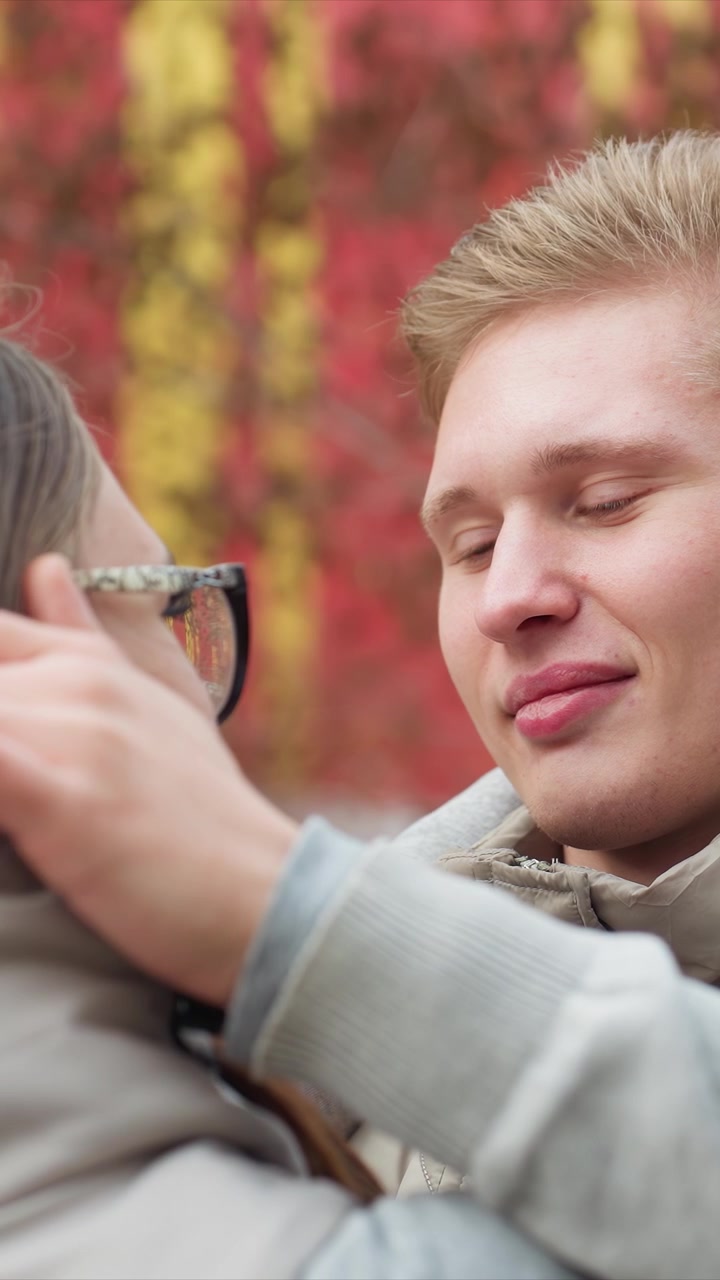 One pair of hands puts or fixes spectacles on other. Gentle contact is made with woman skin near her mouth. Outdoor scene shows red and yellow autumn leaves in background