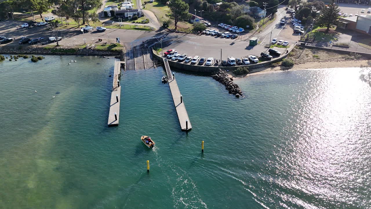 A boat approaches the dock at high tide, navigating through calm waters. The surroundings include a bustling carpark and a scenic landscape, highlighting community activities.