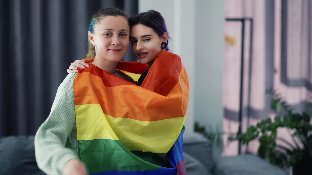 Cheerful women wrapped in colorful rainbow flag at home
