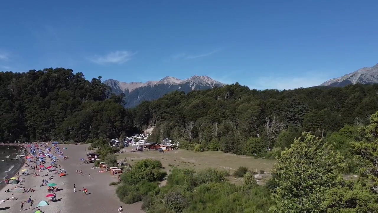 Ripresa aerea con carrello laterale da una spiaggia turistica a Villa La Angostura, con la catena montuosa delle Ande sullo sfondo in una giornata di sole.