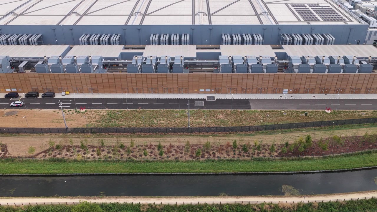 Moderne datacenterfacilitet set ovenfra om dagen med biler, der kører langs en nærliggende vej, og et grønt landskab med en kanal i det omkringliggende industriparkområde.