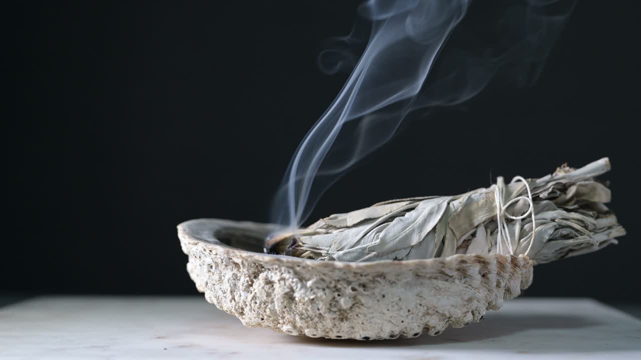 Burning sage in a shell on a wood table with black background showing sage smoke
