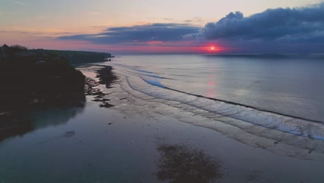 fixed drone clip over calm beach in bali, indonesia with gentle rolling waves lapping onto shoreline, still ocean and pink sunset