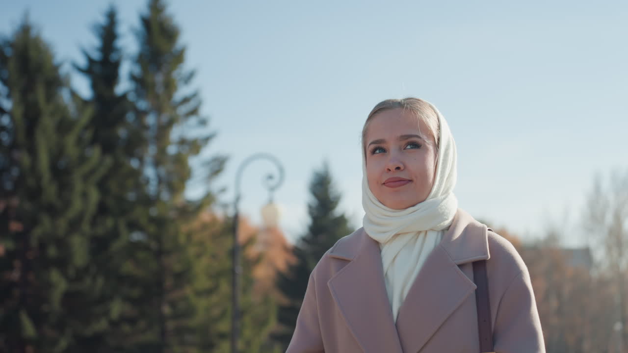 Белая женщина улыбается на фоне сосен и чистого голубого неба. Крупный план лица в шарфе и пальто, залитый осенним солнечным светом, мягкий вязаный шарф, легкий ветерок, довольное выражение лица. Модный портрет с естественным фоном.