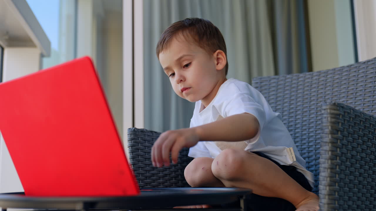 Little cheerful curious baby boy sits in the chair. Cute kid plays on the laptop at home. Low angle view