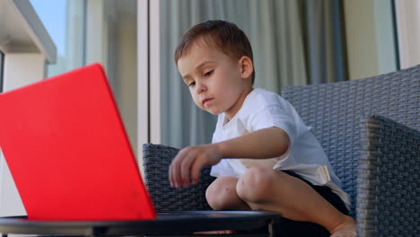 little cheerful curious baby boy sits in the chair. cute kid plays on the laptop at home. low angle view
