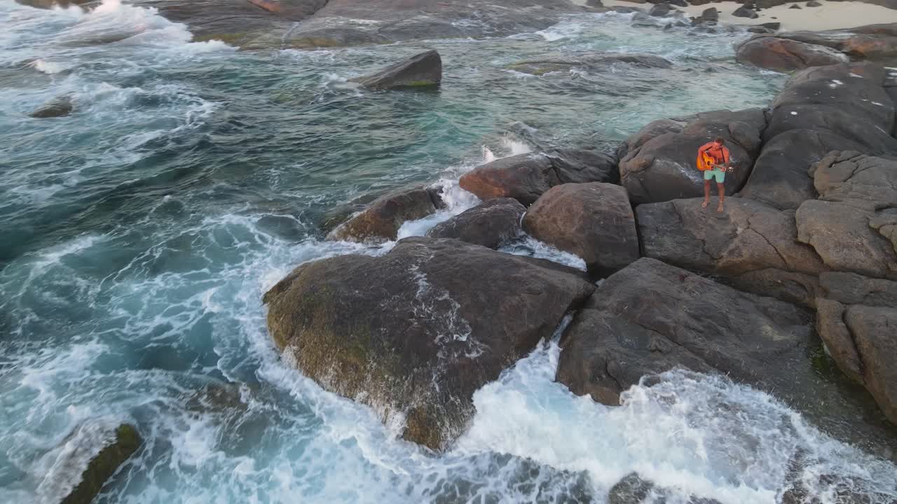 4k increíble tiro aéreo atractivo joven tocando la guitarra en las rocas frente a las olas del mar salvaje al atardecer estilo de vida lento 2