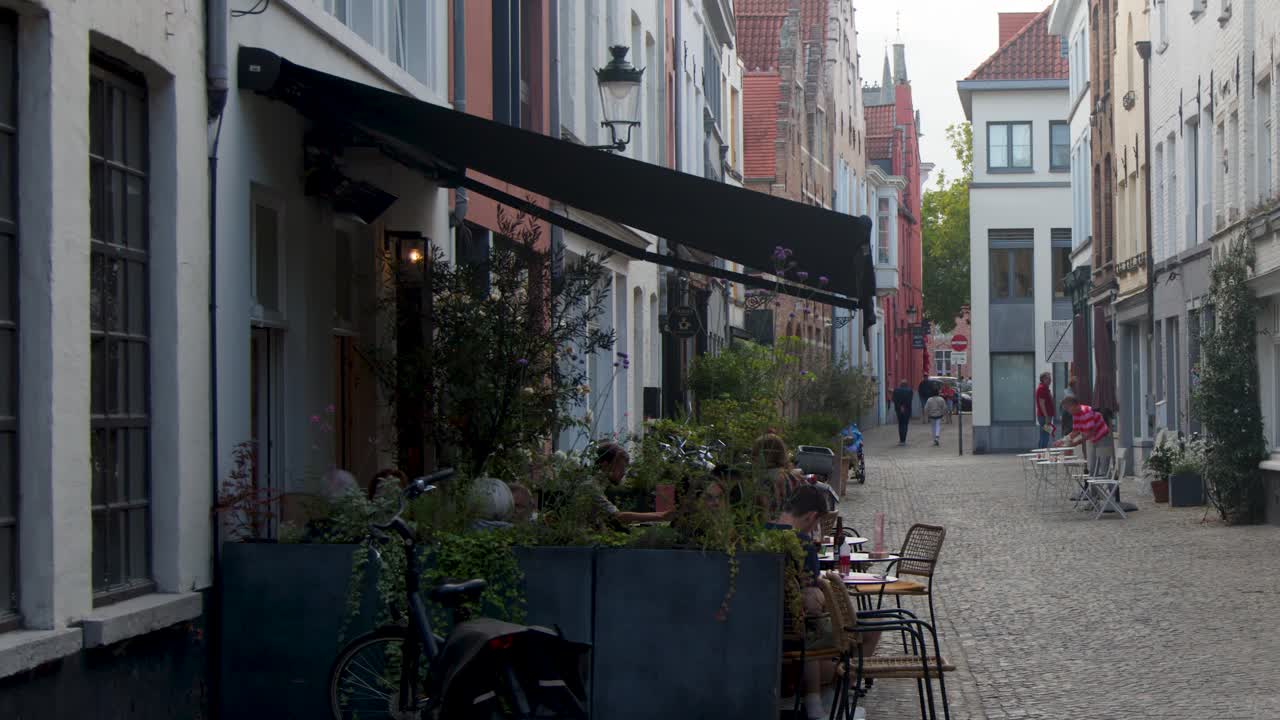 People walk along a cozy cobblestone alley lined with outdoor cafes and historic buildings
