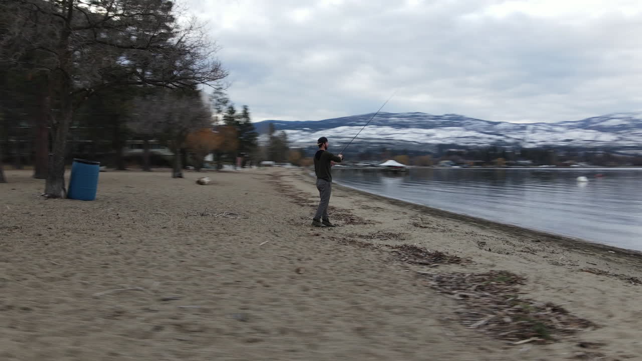 vista aérea de un joven con camino de pesca en la costa del lago okanagan, kelowna, columbia británica, canadá con colinas cubiertas de nieve en el horizonte, disparo de drone en órbita