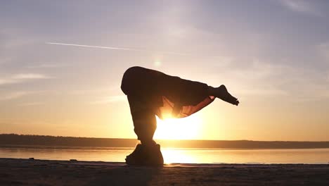 the girl does asana in yoga, slowly raises her legs in the rack on her head. silhouette of a woman in the background of the shining sun