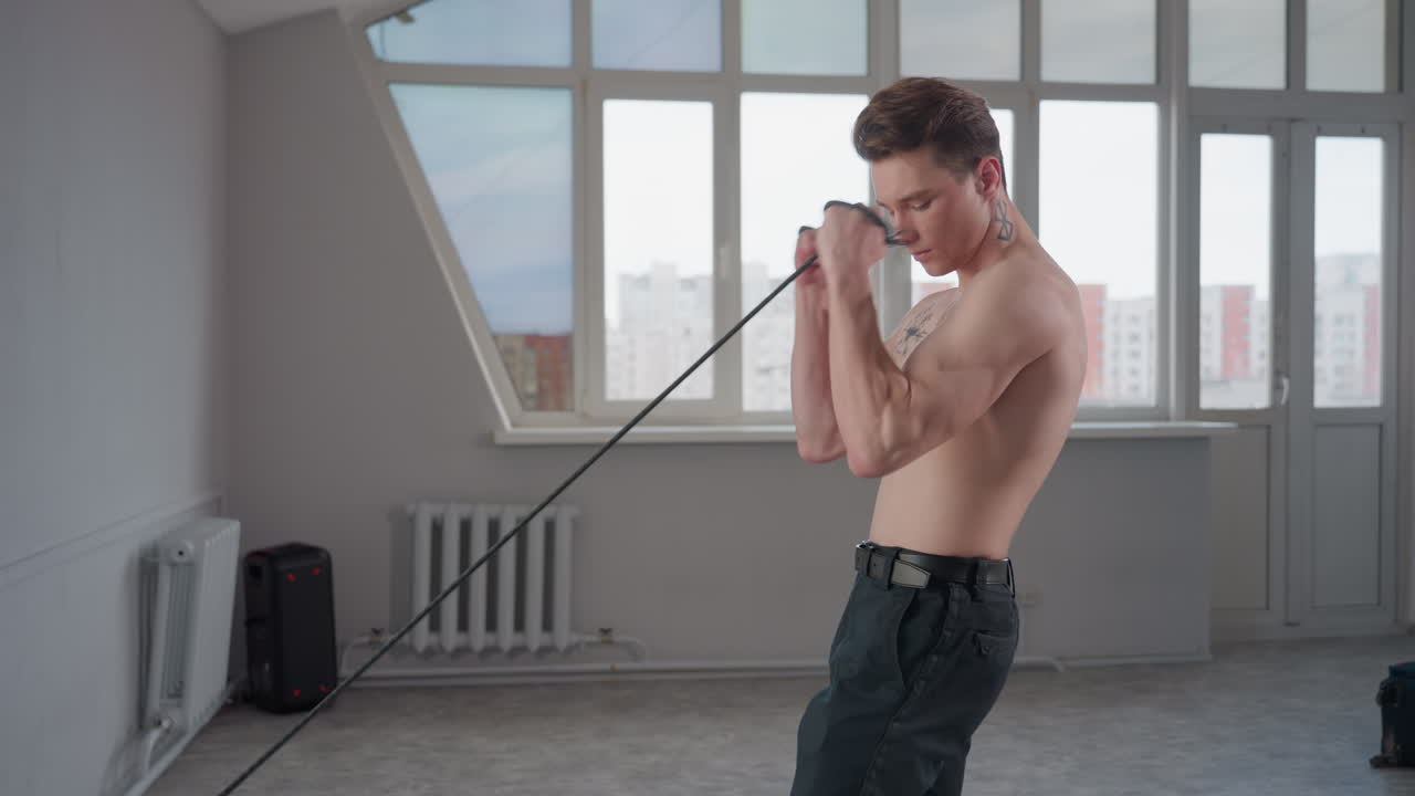 teen shirtless with geometric neck tattoo stands in bright studio by large window, grips heavy rope and pulls with full body effort, demonstrating strength training