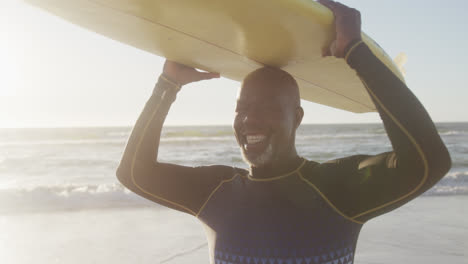 Retrato-De-Un-Feliz-Hombre-Afroamericano-Mayor-Sosteniendo-Una-Tabla-De-Surf-En-La-Playa,-En-Cámara-Lenta