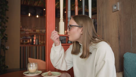 side view of woman with glasses wearing white sweater enjoying dessert with toothpick while seated at round table in cozy cafe with warm lighting and latte on saucer beside dessert plate