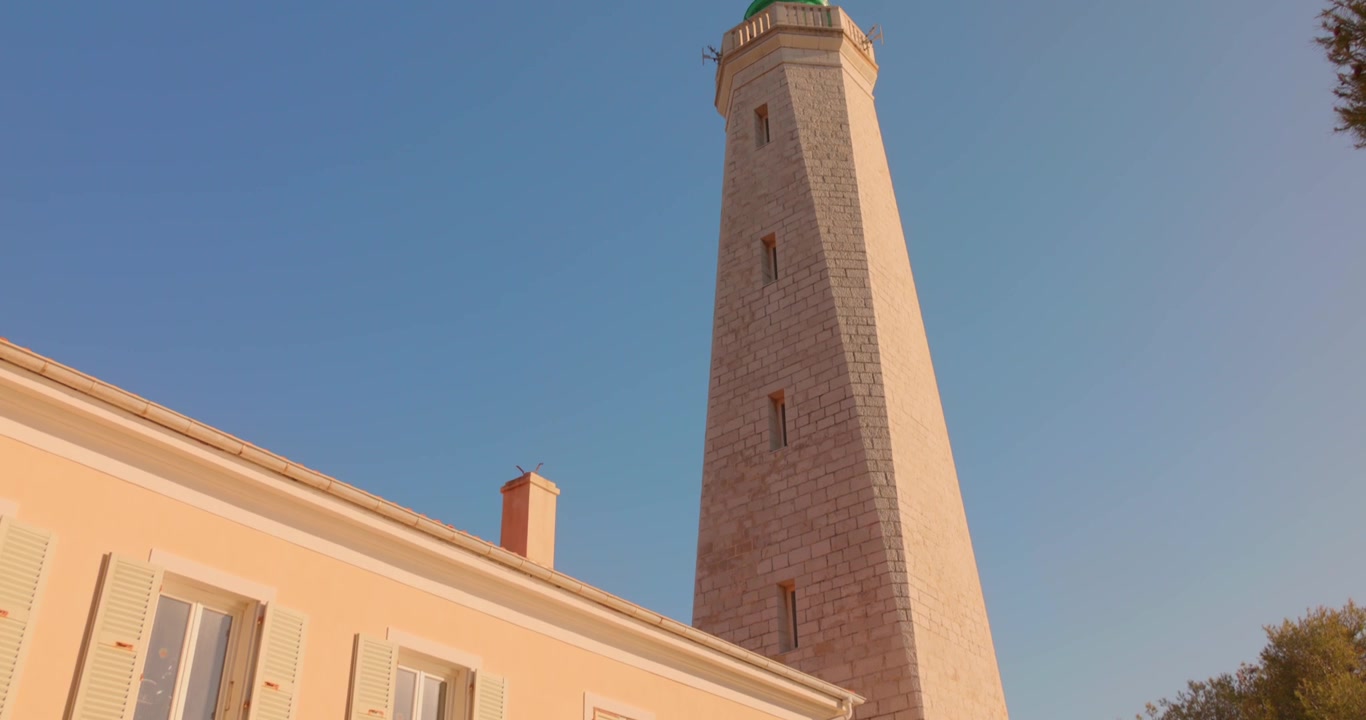 Vista dal basso di un faro e di un edificio adiacente sotto un cielo azzurro limpido alla luce del sole.