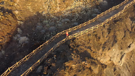 mujer con un vestido rojo camina por un camino con una barandilla en una isla de lava al atardecer dorado, charco de los sargos - disparo de drones