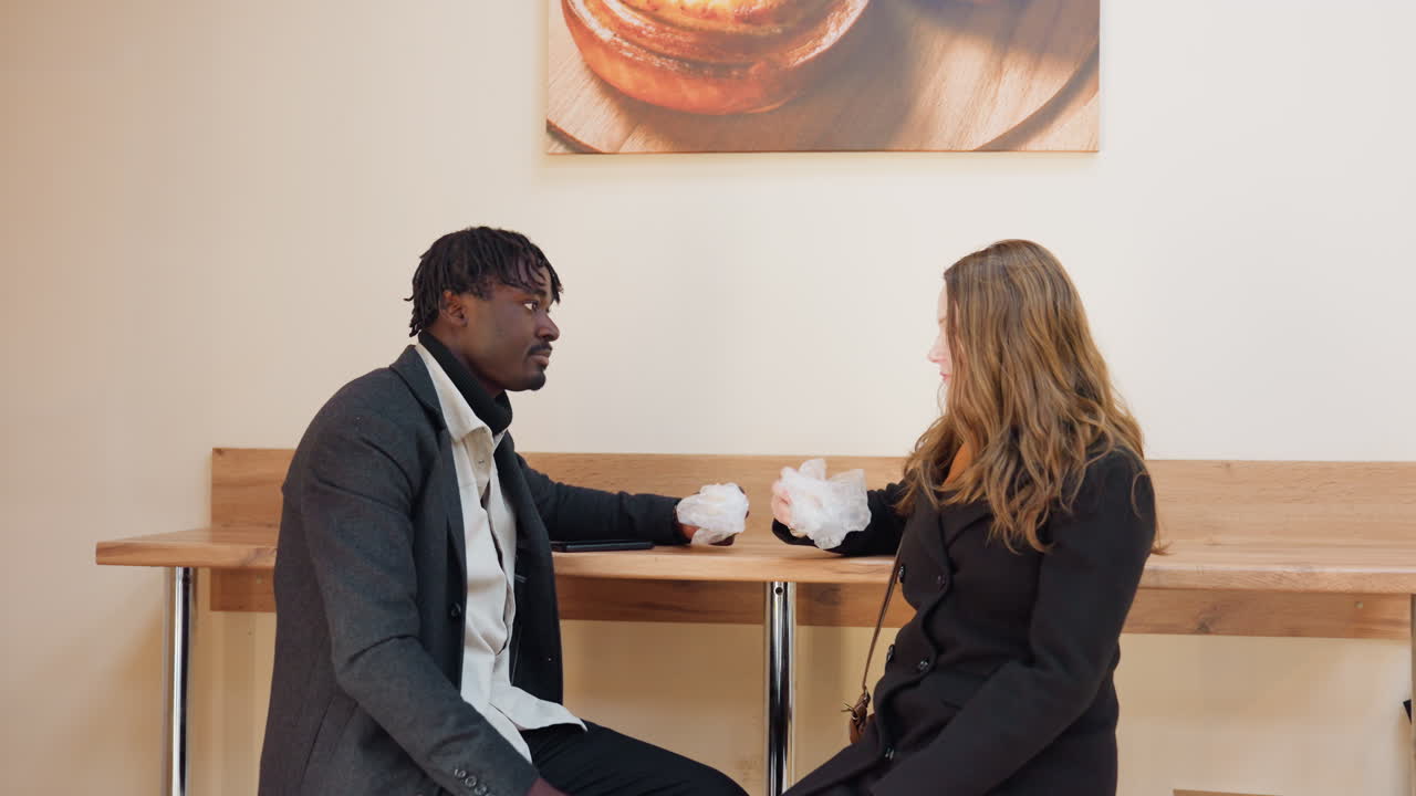 Two friends sitting together eating snacks in calm indoor setting, sharing relaxed moment with friendly interaction and natural expressions, dressed warmly while enjoying food and conversation t