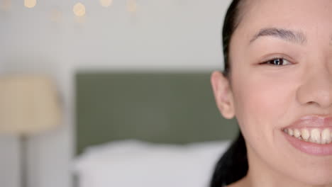 Close-up-of-a-young-biracial-woman-smiling,-with-a-blurred-bedroom-background,-with-copy-space