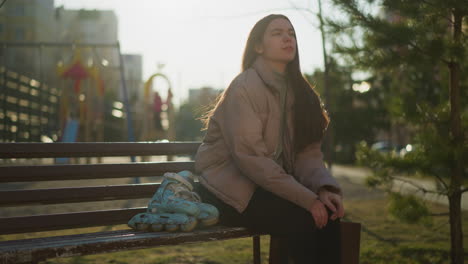 young girl wearing a peach jacket, grey inner shirt, and black trousers sits on a park bench with her rollerblades beside her. she rests one hand on her chin in deep thought, with a blurred background