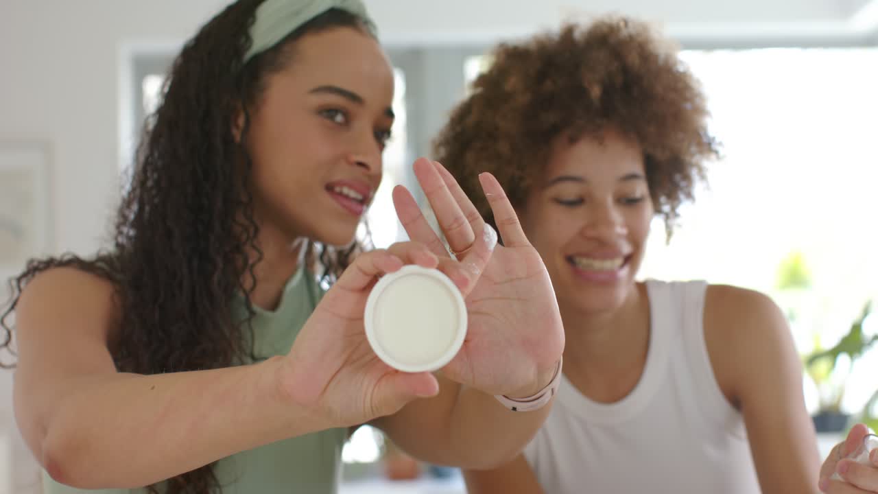 Two African women showing cream in kitchen, left extending jar, right pumping, both testing texture