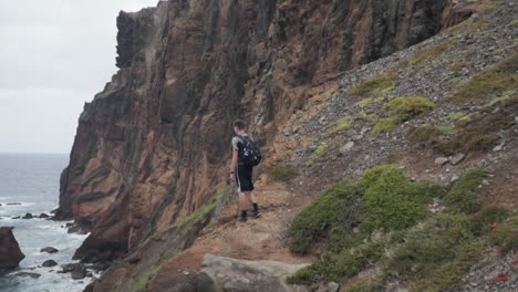 Man-stands-on-rocky-bluff-overlooking-ocean-and-shoreline,-slow-motion