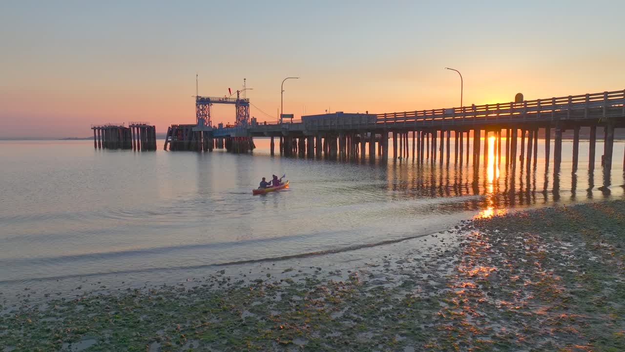 una pareja joven despega y rema juntos en kayak de mar al amanecer flotando bajo el muelle con el resplandor del sol reflejándose en el agua de la bahía cerca de la vista aérea de drones de seattle, washington