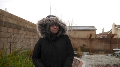 a woman wearing a winter rain coat and hood walking in her garden in a freezing cold winter snow and hail storm slow motion