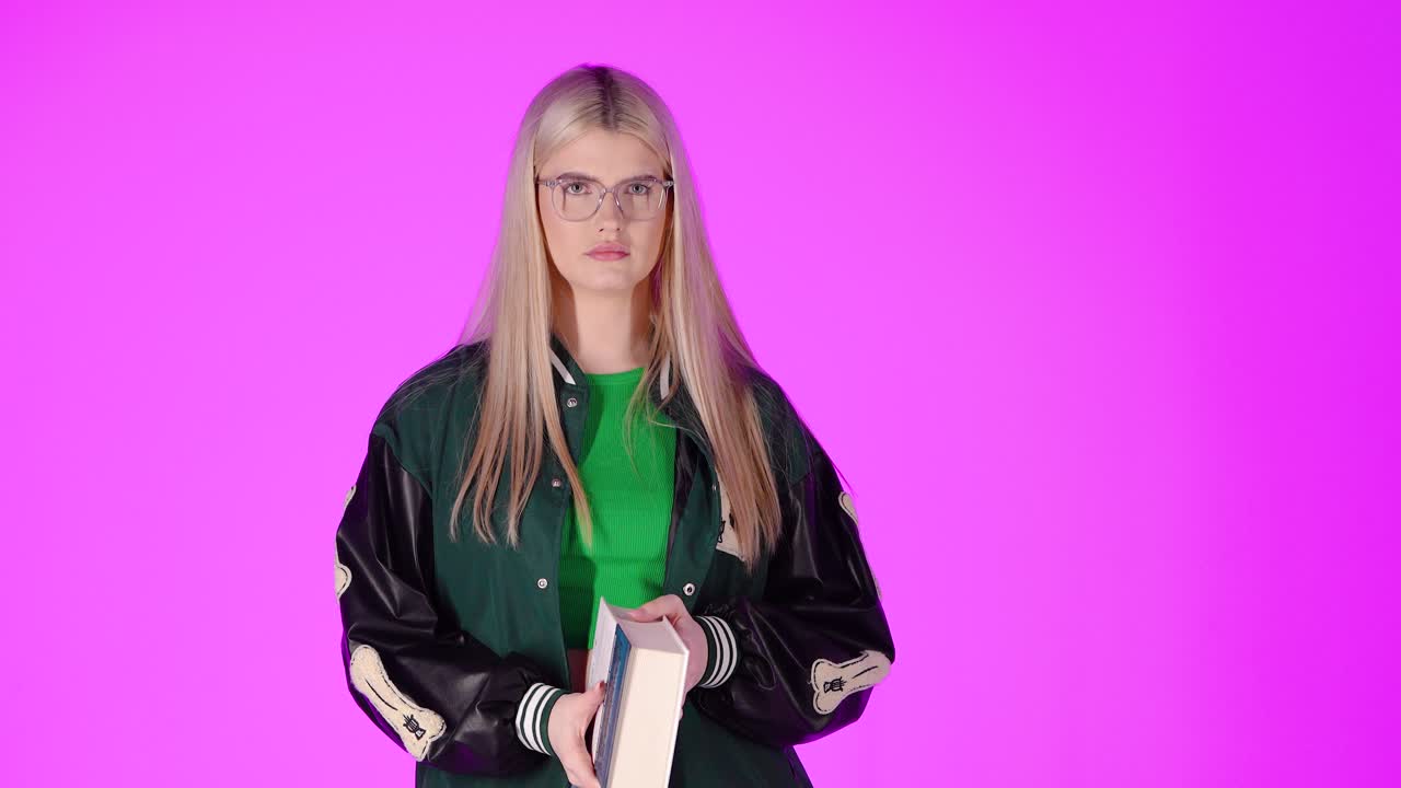 joven estudiante rubia disfrutando de leer un libro y sonriendo, retrato de estudio con fondo rosa