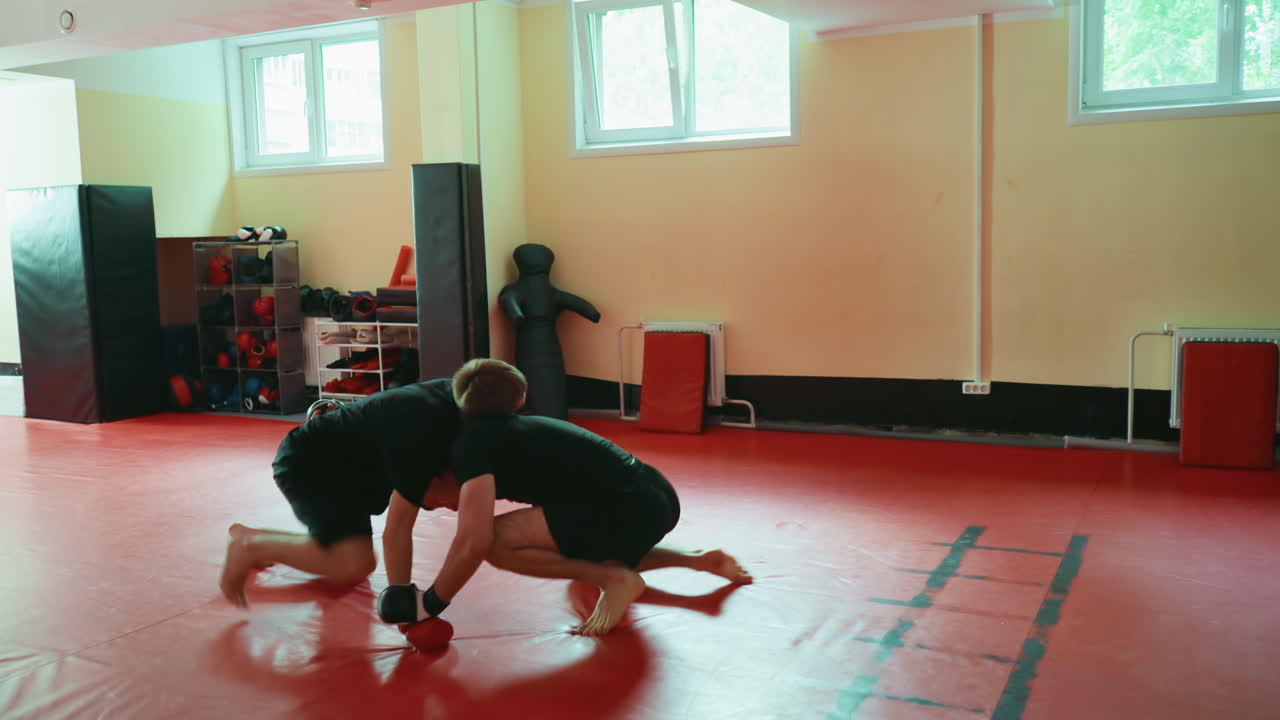 Two male fighters practicing martial arts in gym performing grappling technique as one lifts opponent off red mat floor during sparring training session under window light with equipment in background